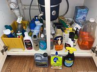 Front view of the under-sink cabinet with various household cleaning supplies including spray bottles, liquid detergents, and cleaning creams, along with plastic wrap boxes.