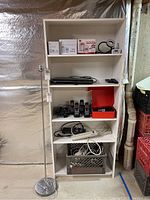 Photo of the white shelving unit containing a silver metal floor lamp stand without shade on the left side, Panasonic cordless phone system with two handsets and charging base, various cords, power strips, and miscellaneous electronics on shelves.