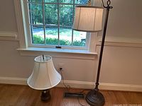 Photo showing the table lamp with a white bell-shaped shade and the floor lamp with a drum-shaped shade near a window on hardwood floor.