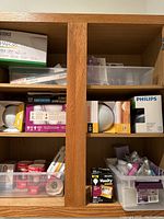 Wide view of wood cabinet with multiple boxes and containers of light bulbs including Philips, Sylvania, and others, some loose, some boxed.