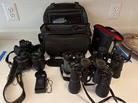 Photo showing Nikon D3200 camera body, two zoom lenses, battery charger, and three binocular sets with cases.