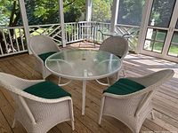 Full view of round glass top outdoor table with four white wicker chairs with green cushions on wooden deck