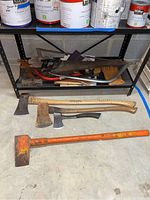 Photo of axes, splitting maul, hand saws, bow saw, and work gloves arranged on and below a metal shelf in basement setting.