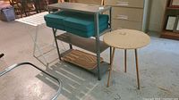 Wide view showing metal shelving unit with teal cushions and wooden mud guard, white folding table, round cork-top table