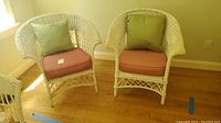 Two white wicker chairs with red seat cushions and green back cushions placed side by side on hardwood floor.