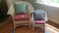 Both adult and child white wicker rocking chairs with cushions and pillows beside a window on wooden floor.