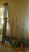 Photo showing a variety of yard tools leaning against a wall, including stakes, rakes, shovels, snow shovels, hole digger, spade, broom.