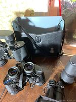 Two Bushnell binoculars, one Tasco binocular, carrying case and binocular pouch displayed on a table.