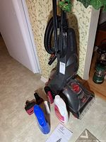 Full view of Bissell upright carpet cleaner with hose wrapped around handle, multiple cleaning attachments, bottles of cleaning solution, and user guide manual on floor nearby.