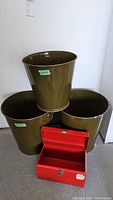 Three olive green metal buckets arranged with one small bucket on top of two larger buckets plus one red metal toolbox open showing empty interior, on concrete floor.
