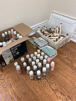 Several acrylic paint bottles arranged upright and laid flat on the floor along with a container holding many tubes of Mayco wood stain paint and some plastic paint mixing palettes.