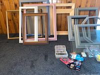 Photo showing various picture frames in different sizes and colors along with picture hanging supplies on a carpeted floor against wood panel wall.