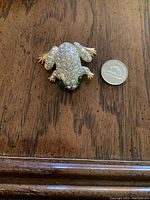 Top view of frog-shaped brooch showing clear rhinestones, green eyes, and gold-tone feet beside a coin for size.