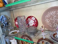 Shelf of assorted glass and ceramic items including crystal vases, decorative plates, and a glass hummingbird feeder shaped like a bird