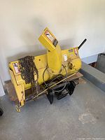Full view of yellow snow thrower attachment showing auger, chute and mounting chains