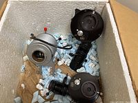 Three microscope components inside a styrofoam container with shock-absorbing packing peanuts, showing the Wild Heerbrugg black accessories and the metallic illuminator.