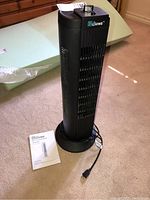 Full view of tall, black oscillating tower fan standing on carpet next to instruction manual and power cord visible.