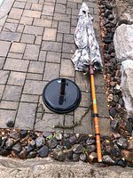 Overview of folded patio umbrella lying next to circular cast iron stand on stone patio with rock border.
