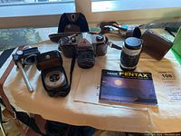 The main collection of items showing the Pentax SLR camera, additional lens, light meter, and manual laid out on a surface.