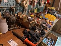 View of vintage bench grinder, electric screwdriver, chargers, and box of roofing nails on workbench with tool pegboard backing.