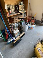 Overview of brooms, manual air pump, white bucket with garden tools, plastic jerry cans, shelving unit with small hardware drawers, cardboard boxes, and various fluids and spray cans