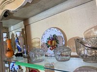 Shelf display of assorted glassware including vases, bowls, and decorative plate with fruit illustration.