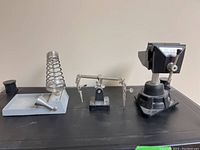 Three items shown on a black surface: soldering iron holder with coil spring on the left, adjustable clip holder with articulated arms and clips in the center, and D-vice by Dremel 23075 vise with rotating base on the right.