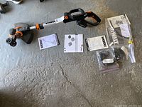 View of the full Worx weed trimmer laid on floor with manuals and instruction leaflets spread alongside it showing included documentation and components.
