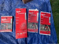 Four red boxes of Milwaukee bandsaw blades arranged on a blue tarp, unopened packaging visible.