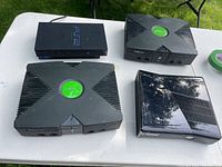 Four game consoles on white table: Sony PS2, two Original Xbox consoles, and one Xbox 360 console