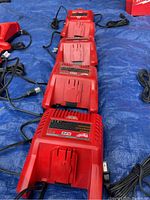 Five Milwaukee M28 battery chargers lined up on blue tarp with cords unplugged, showing front side and battery connection slot.