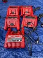Seven red Milwaukee M18 battery chargers shown on blue tarp, arranged with cords visible.