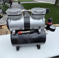 Photograph of the airbrush compressor showing the dual-cylinder motor mounted on a black tank, pressure regulator knob with moisture trap attached.