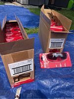 Two open cardboard boxes full of individual Milwaukee tinted safety glasses packages. One pair is placed outside the boxes for display, showing red packaging and black frame glasses with tinted lenses.