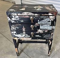 Front view showing three drawer nightstand with black lacquer and painted figures, metal ornate drawer pulls, and paint chipping on legs.