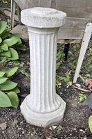 Front and side views of a light gray concrete garden column pedestal with vertical grooves and octagonal base and top, placed outdoors in garden soil among plants.