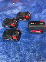 Four Milwaukee M18 battery packs arranged on a blue tarp (three standard, one XC8)