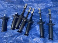 Seven new black Milwaukee tool handles and grips arranged on blue tarp, showing side and top views with mounting attachments visible.