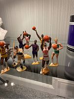 Collection of assorted sports figurines, mostly basketball players including Michael Jordan, Magic Johnson, Kareem Abdul-Jabbar, Larry Bird, and Isiah Thomas, plus a baseball figurine and a VHS tape visible