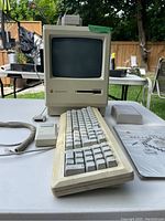 Front view of Apple Macintosh Plus computer, keyboard, mouse, and manuals on table outdoors.