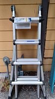Front view of folded aluminum ladder leaning against a wall with instruction manual attached.