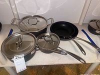 Several stainless steel pots with glass lids and non-stick frying pans arranged on a shelf surface.