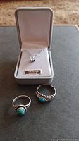 Sterling silver necklace displayed in gray box, accompanied by two rings with turquoise stones on dark surface