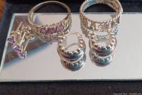 Display of three sterling silver rings and two pairs of earrings on a mirrored surface.