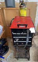 Photo of wire feed welder showing red and black metal case with control panel and attached hose and cable, positioned in front of wooden panel and other items.