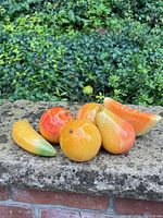 Front view of six alabaster carved stone fruit pieces placed on a stone ledge outdoors with greenery in the background.