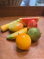 Seven pieces of carved and polished alabaster fruit including watermelon slice, apple, lemon, pear, and others, placed on a wooden surface, showing their detailed coloring and natural stone veining.