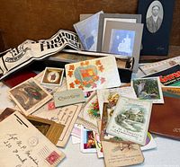 Assorted ephemera spread including fold-out cloth banner, mounted black and white photographs, various cards, and postcards with colorful illustrations.