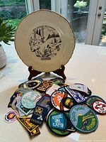 Front view of the ceramic Boy Scouts of Canada camping plate on stand with a large dozen embroidered patches in front