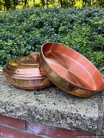 Copper pot with wood handle and lid beside the round copper baking pan on a stone ledge outdoors.